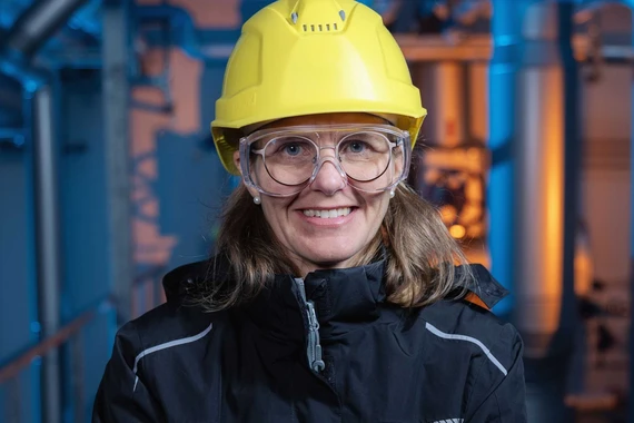 Frau mit verschränkten Armen, Schutzbrille und gelbem Helm in industrieller Umgebung