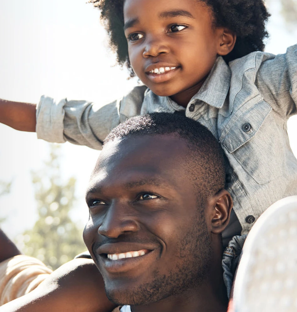 Foto de um homem sorrindo com uma criança sentada sob os seus ombros de braços erguidos para o ar, ao fundo da imagem aparece o céu e os galhos de árvores, com folhas. O homem é adulto, preto, de cabelo curto. A criança é preta, tem cabelos cacheados médio escuro, usa uma blusa social e calça jeans. Ambos olham para o horizonte num dia ensolarado. 