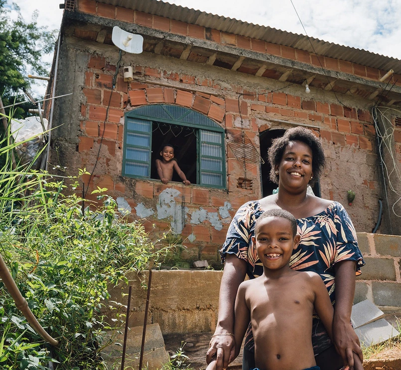 Foto de uma mulher abraçando uma criança por trás, ambos sorriem para a câmera. A mulher é negra e a criança também. Atrás de ambos, uma casa rural de tijolos com uma criança sorrindo através da janela. Ao redor da casa estão várias árvores.