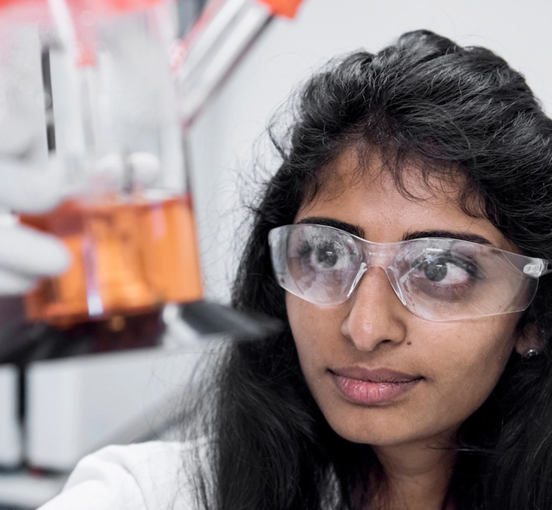Foto de uma pessoa com óculos de proteção segura um objeto desfocado no ar. A pessoa é uma mulher, morena, com cabelo liso preto. O fundo é desfocado e tem paredes brancas. 