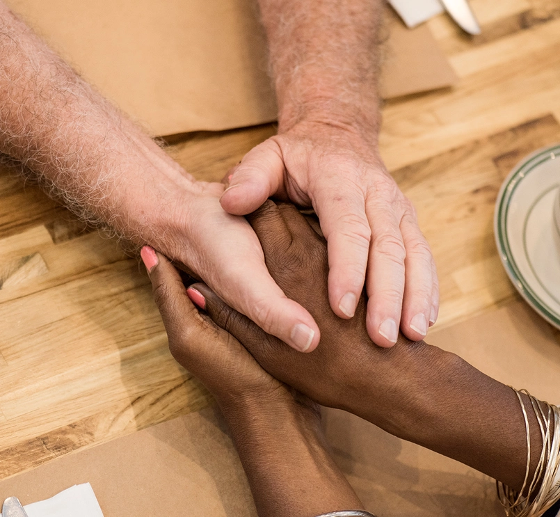 Foto em plano detalhe da vista de cima de uma mesa com xícara de café. Ao centro, duas mãos abraçam a mão de outra pessoa. 