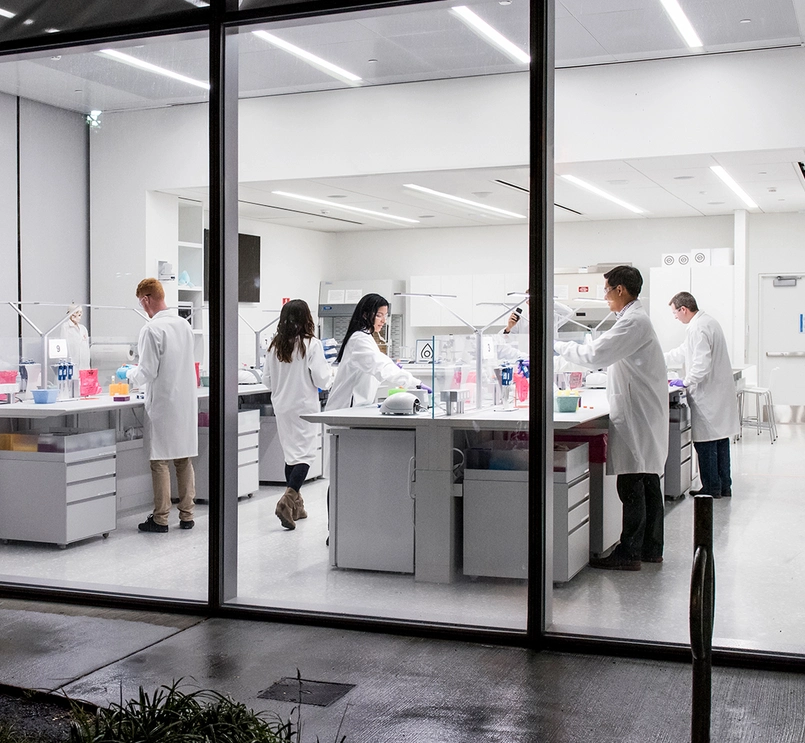 Foto de uma sala de laboratório vista através das janelas altas. O laboratório é iluminado com luz branca, com bancadas, chão e paredes brancas. Nas bancadas estão frascos coloridos e adjacente mulheres e homens de jaleco branco trabalham.