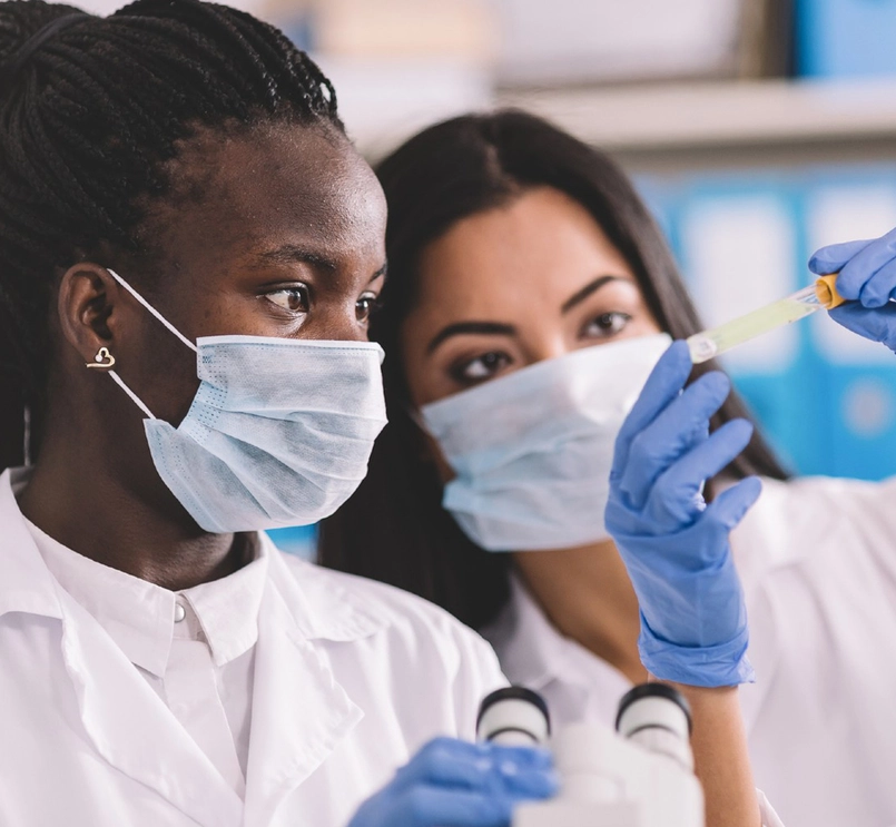 Foto de duas pessoas usando máscara cirúrgica e roupa branca. Uma delas segura uma âmpola amarela usando luvas. Ambas observam a âmpola. 