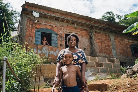 Foto de uma mulher abraçando uma criança por trás, ambos sorriem para a câmera. A mulher é negra e a criança também. Atrás de ambos, uma casa rural de tijolos com uma criança sorrindo através da janela. Ao redor da casa estão várias árvores.