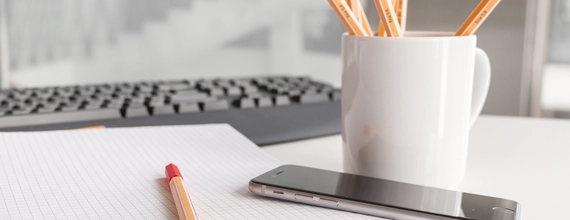 Foto em detalhe da superfície de uma mesa branca com um celular, caneca cheia de canetas de corpo laranja e um teclado preto. 