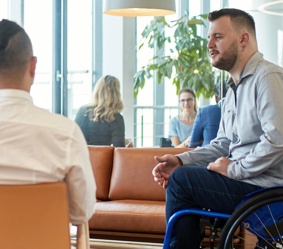 Foto de uma sala com parede de janelas brancas, sofá e poltrona terracota, lustre branco em semi-lua com luz amarela e plantas em vasos no chão. Um homem de blusa branca está sentado na poltrona de costas para a câmera e ao lado dele um homem de cadeira de rodas conversa com ele, vestido de blusa social cinza e calça jeans azul. 