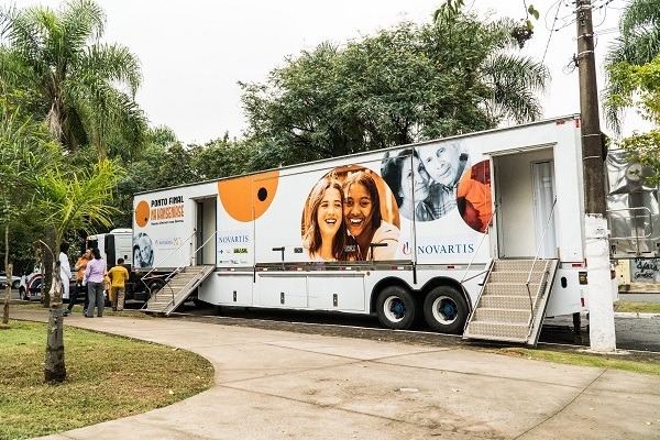 Foto de uma carreta adesivada com logotipo novartis, bolhas laranja e pessoas. As portas estão abertas e algumas pessoas se encontram nas entradas. O ambiente é externo e rodeado de árvores. 