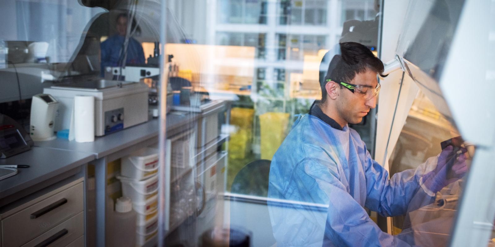 Foto de um homem, moreno, dentro do laboratório, trajado de vestimentas de proteção azul e óculos transparente. Atrás dele, fileiras de maquinário. 