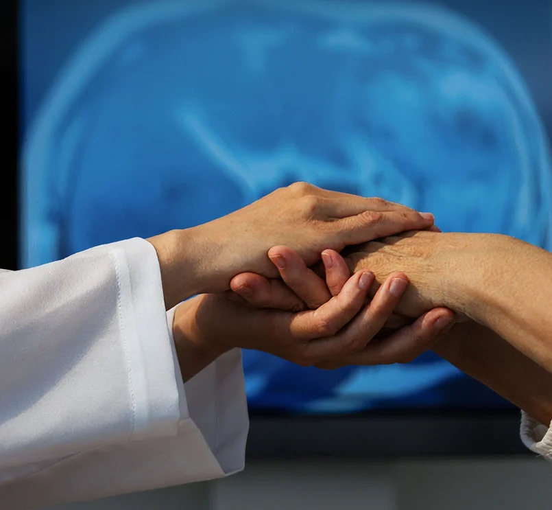 Ärzte untersuchen die Hand eines Patienten, im Hintergrund ein Gehirnscan.