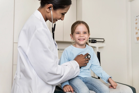 Eine Frau im weißen Kittel horcht ein Mädchen mit einem Stethoskop ab.