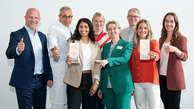 Gruppe lächelnder Personen in Business-Kleidung, mehrere halten Flyer, zwei zeigen den Daumen nach oben