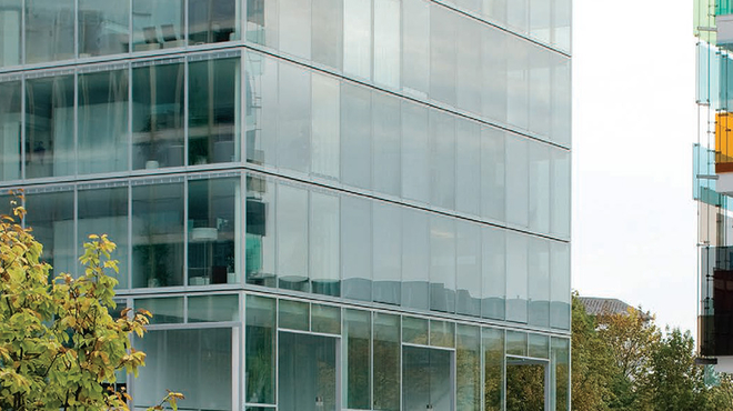 Menschen gehen über einen hellen Platz vor einem Glasgebäude mit Bäumen