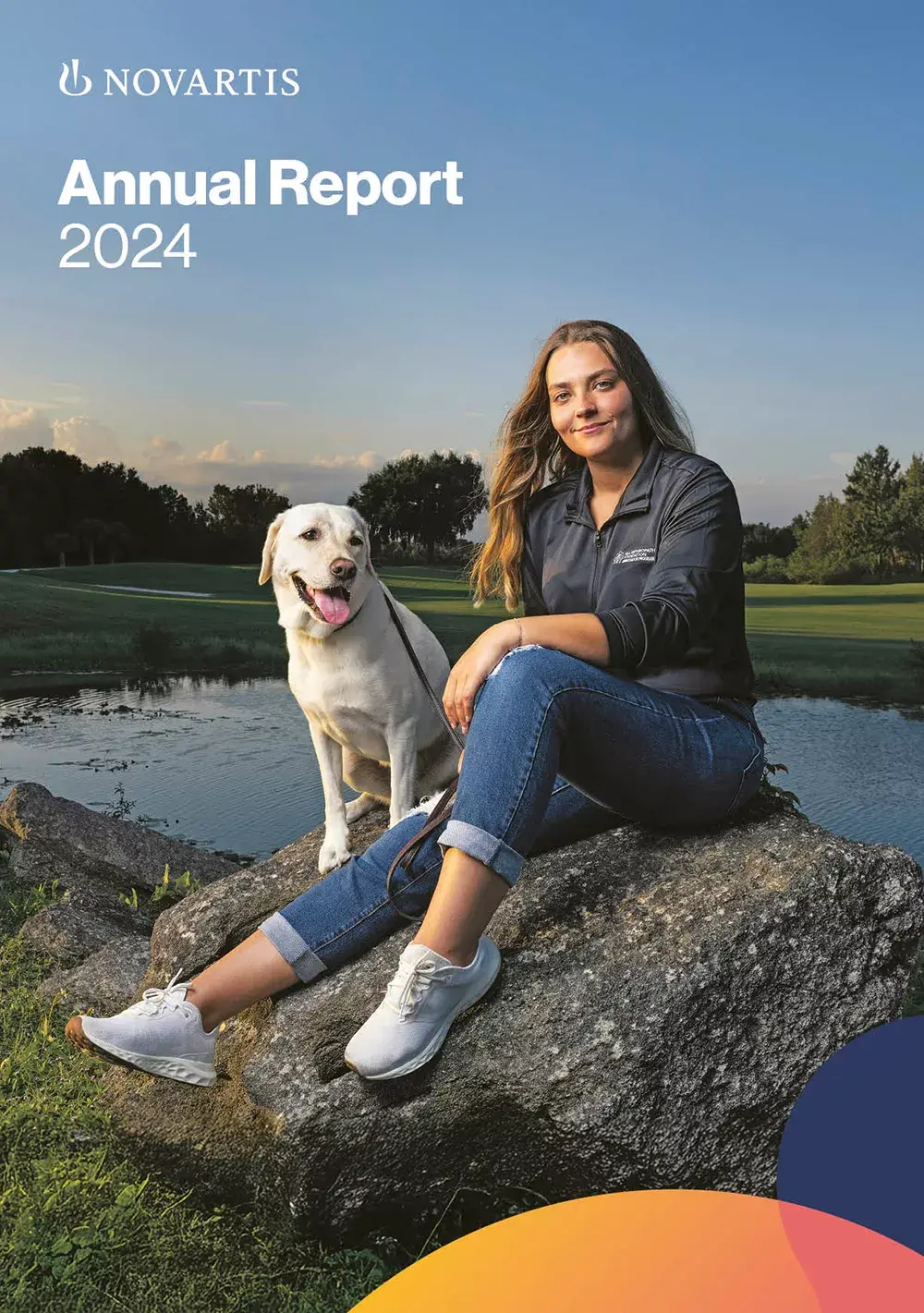 Lächelnde junge Frau in dunkler Arbeitsjacke sitzt neben einem hellen Hund an einem Teich