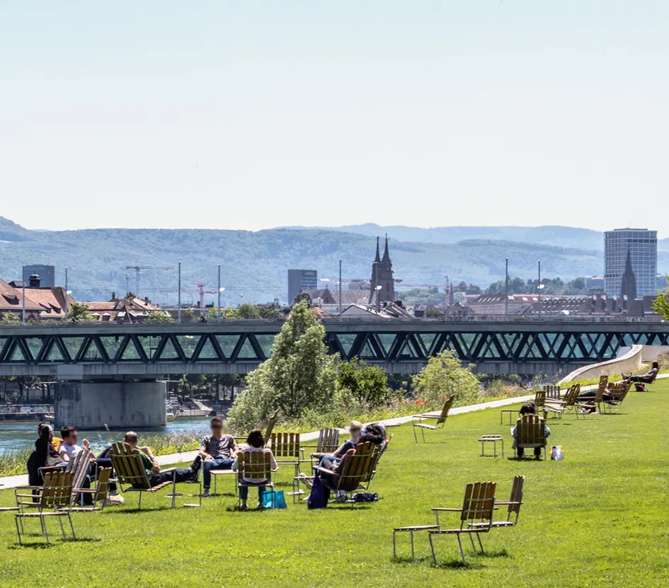 A panoramic view of Basel, from the Novartis Campus with people lounging in the foreground and the black forest in the background