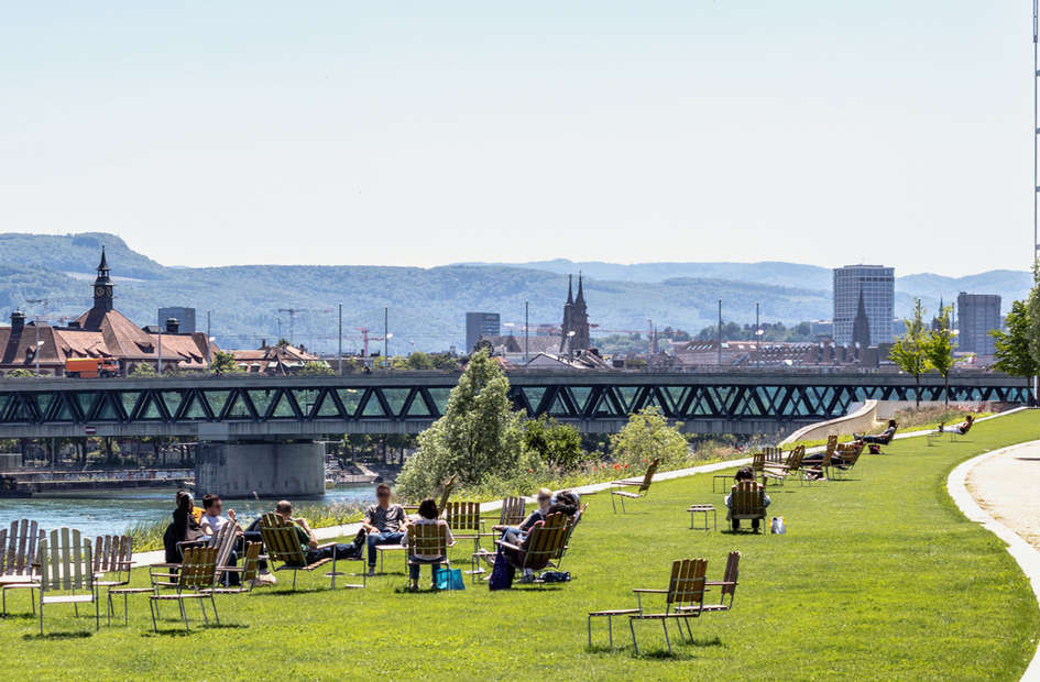 A panoramic view of Basel, from the Novartis Campus with people lounging in the foreground and the black forest in the background