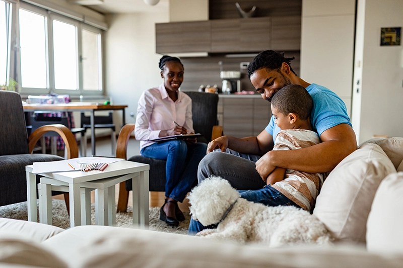 Child hugs dad next to dog on couch, while visiting clinician looks on and smiles