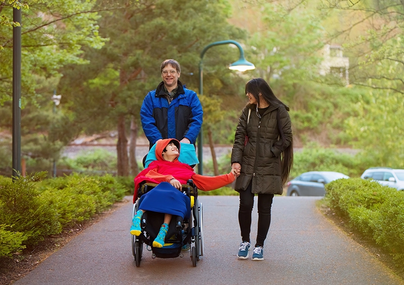 Dad pushes pre-teen boy, wearing red sweatshirt, in wheelchair through park, as boy smiles up at mom. 