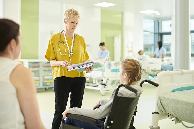 Middle-aged female physician evaluates young girl with braided pigtails in wheelchair, in hospital setting