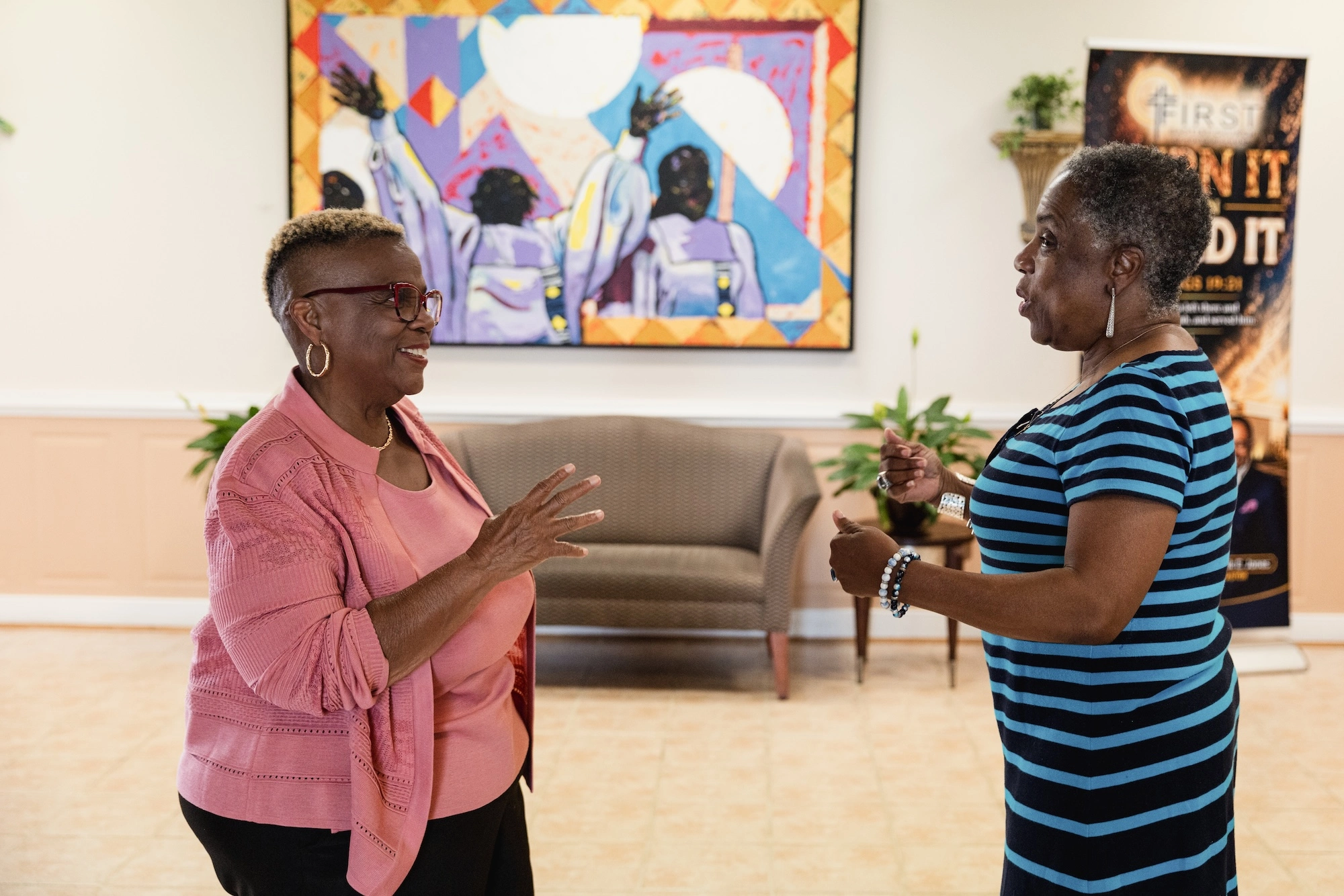 Dr. Pernessa Seele (left) speaks with another attendee in a community meeting space, with a colorful artwork and seating area visible in the background.