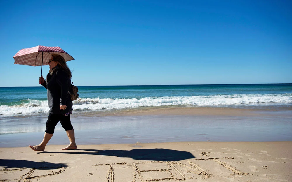 Woman walks along the beach holding an umbrella to protect herself from the sun.