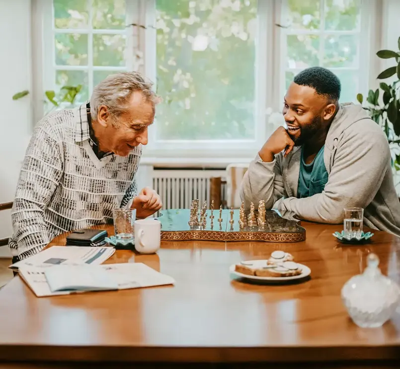 An older man and a younger man playing chess at a table indoors, laughing and engaging in friendly conversation. The room is warmly lit, with plants, newspapers, and refreshments on the table.