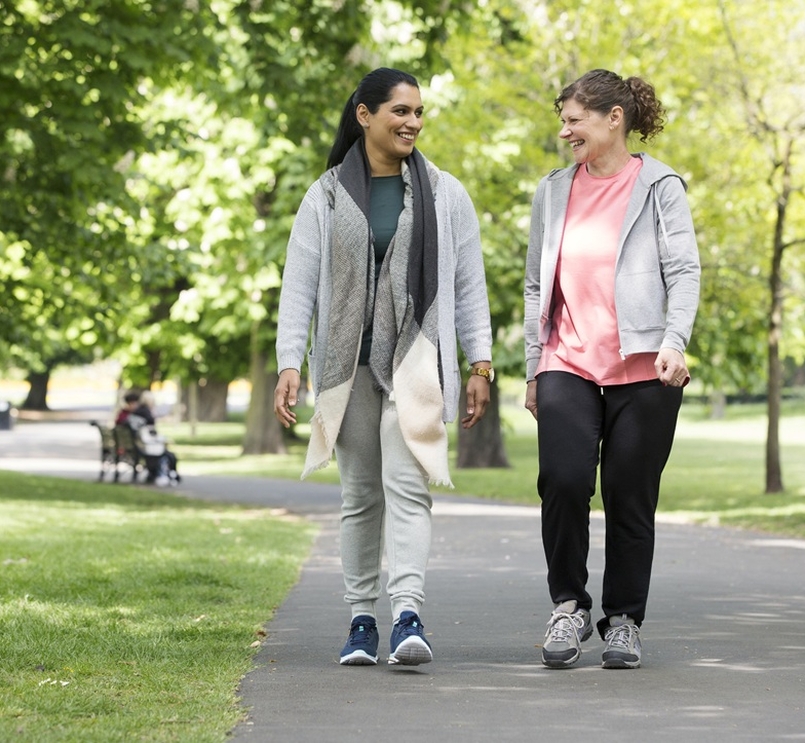 Two women smiling as they walk together through a sunny park, enjoying a friendly moment.  