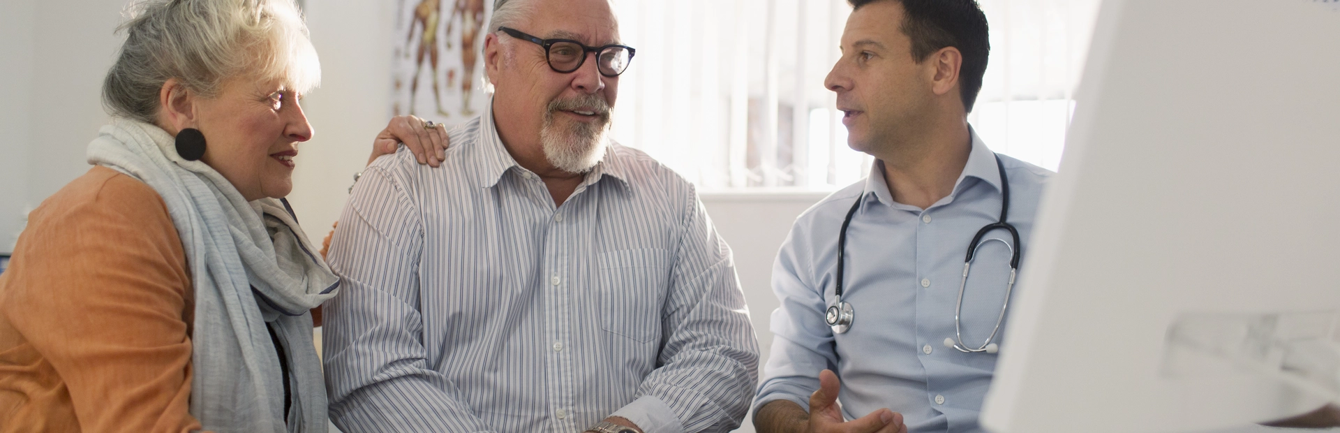 Doctor consulting with older male patient and his wife about prostate cancer treatment options while reviewing information on computer screen in medical office