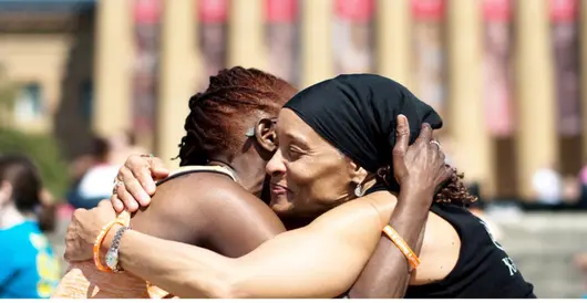 Two smiling women embrace on the steps of a museum, surrounded by event participants in matching shirts. The setting and bright blue sky evokes wellness and community.