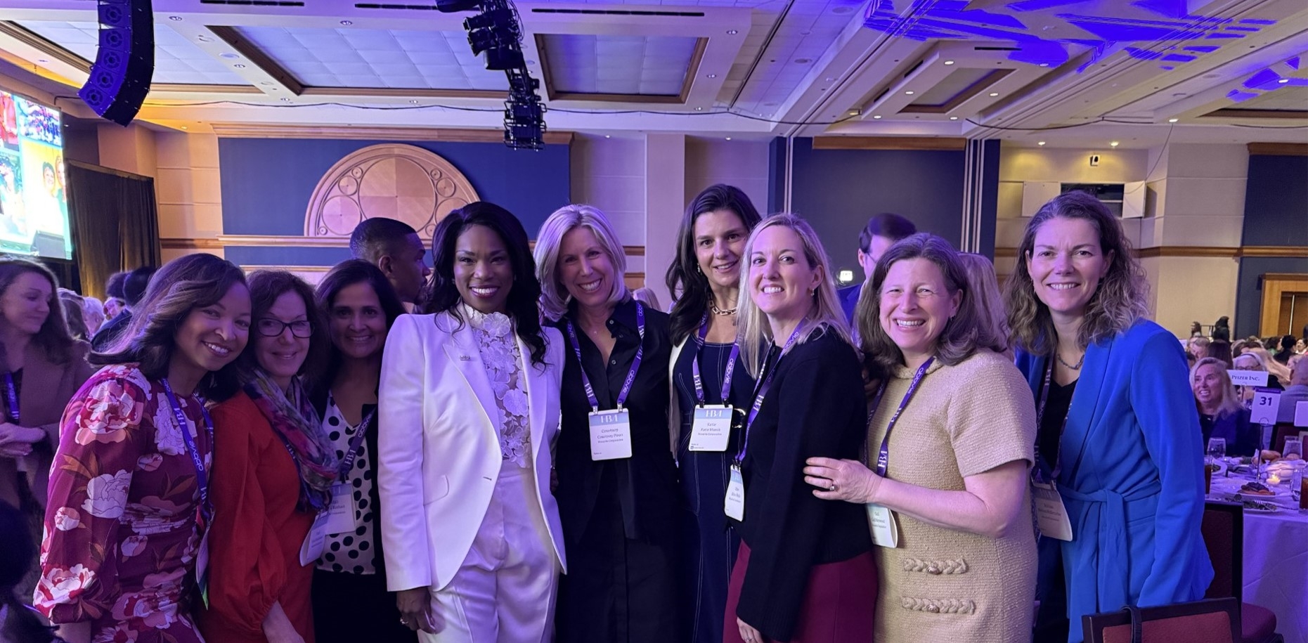 A group of nine women, dressed in business and formal attire, pose together at an event.
