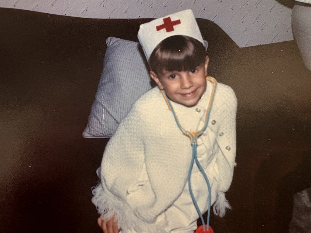 A young child dressed as a doctor, wearing a white hat with a red cross and a toy stethoscope around their neck