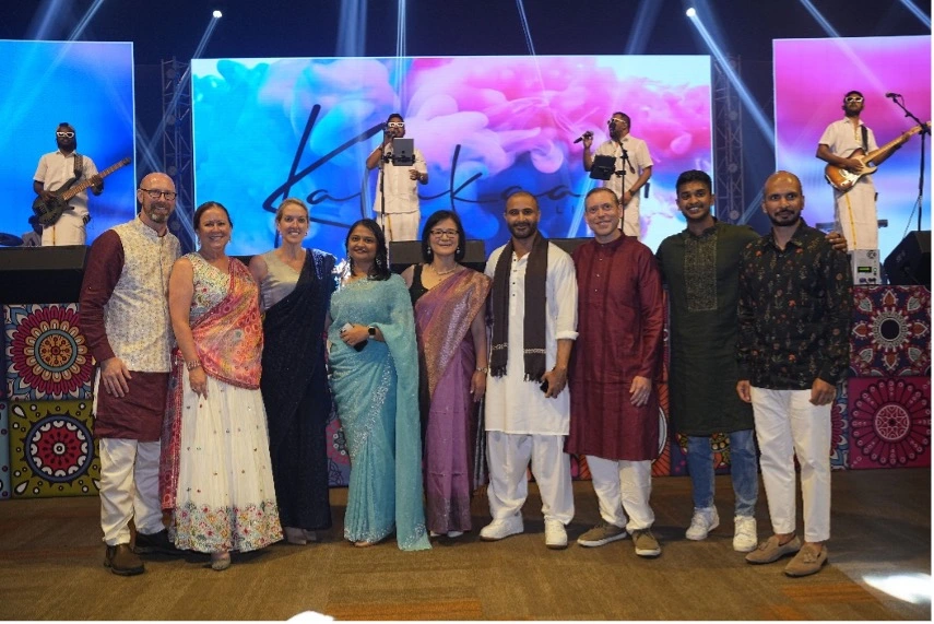 A group of people in colorful attire stand together, smiling. Behind them, musicians perform on stage
