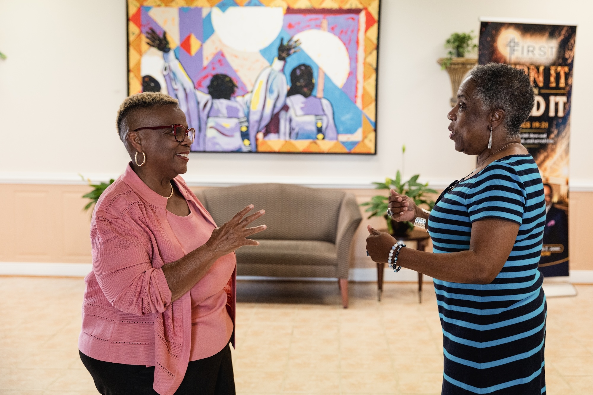 Dr. Pernessa Seele (left) speaks with another attendee in a community meeting space, with a colorful artwork and seating area visible in the background.