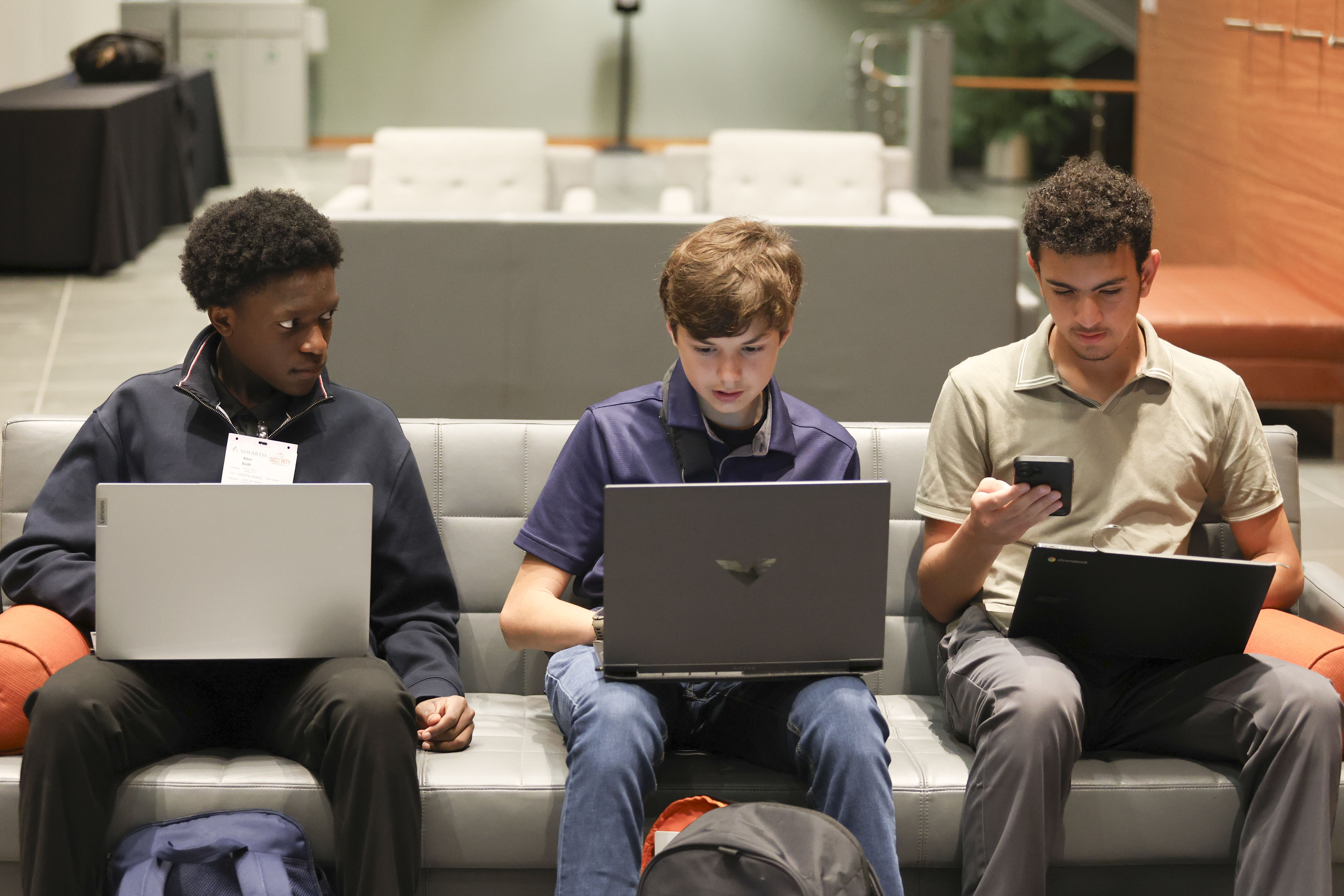 Three people sit on a couch using laptops and a phone, with a basketball resting beside them.