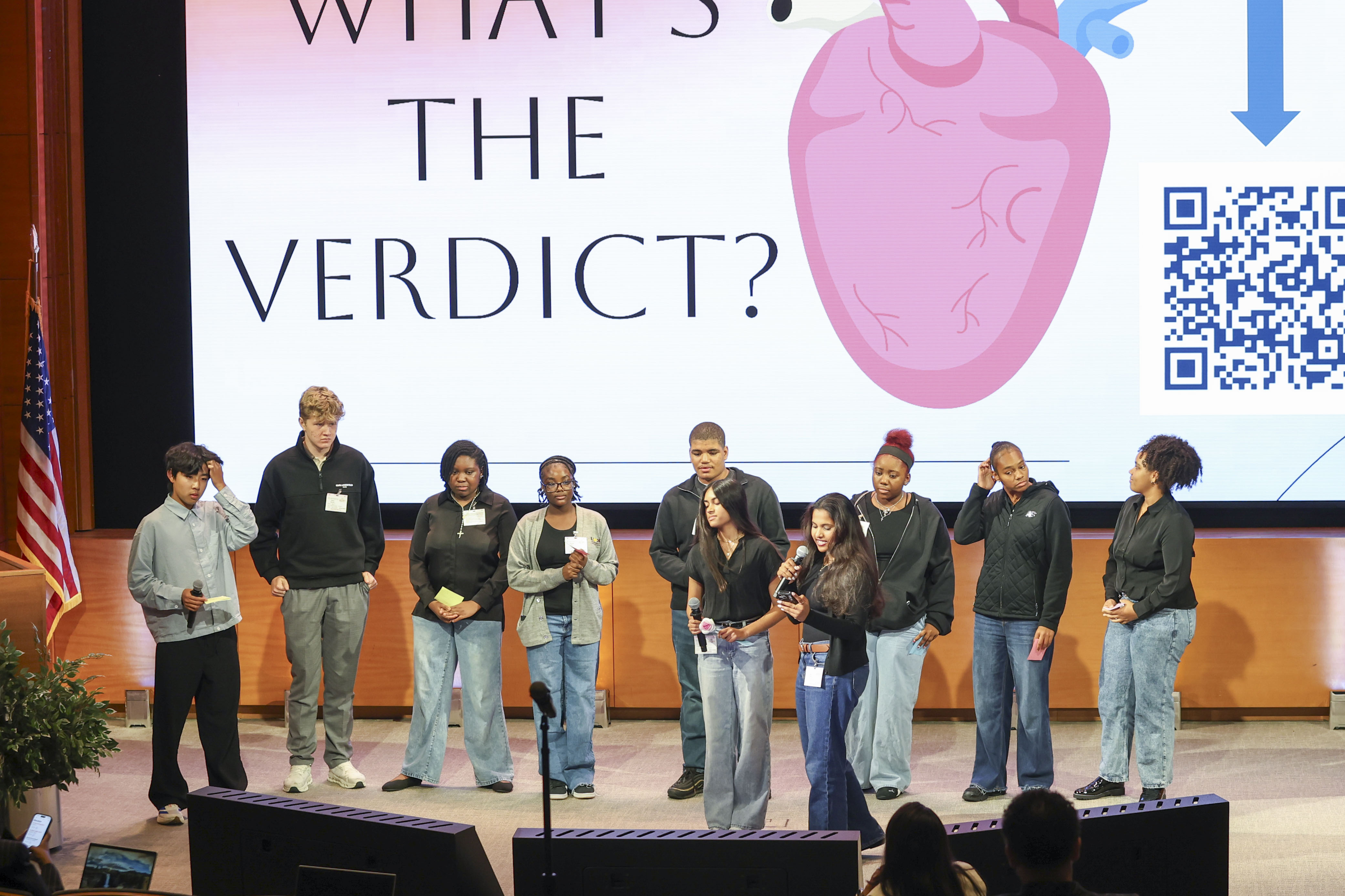 Group of people on a stage in front of a large screen displaying the text “WHAT’S THE VERDICT?”